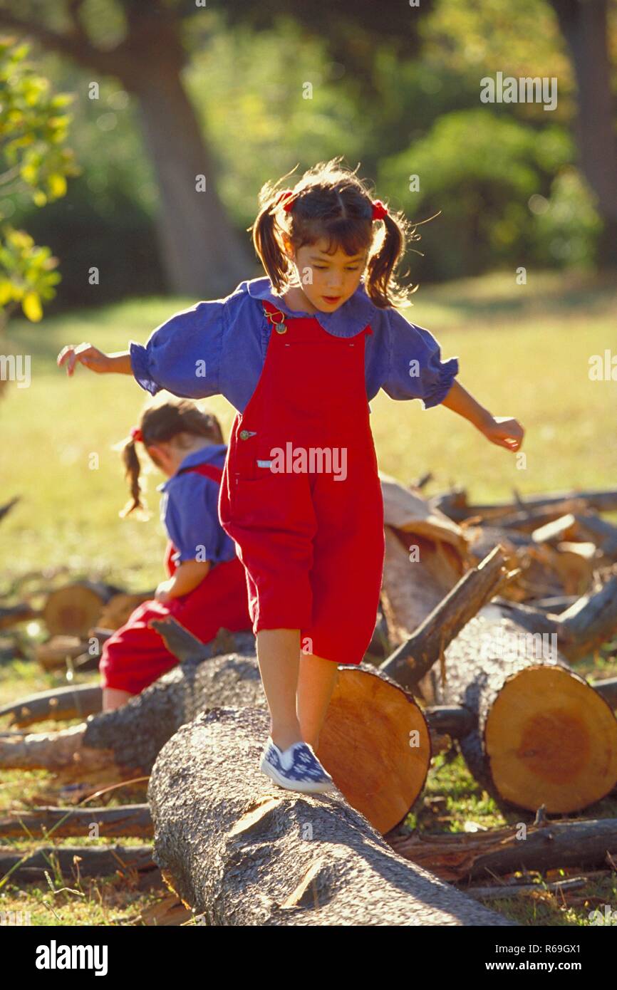 Parkszene, Ganzfigur, 2 bruenette Zwillings-Maedchen, 6 Jahre alt, bekleidet mit roter Latzhose, blauer Bluse und Turnschuhen balancieren ueber dicke Baumstaemme Stock Photo