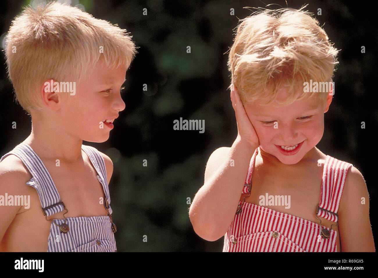 Portrait, Nahaufnahme, zwei 4 Jahre alte Zwillings-Jungen mit kurzen blonden Haaren bekleidet mit blau und rot-weiss gestreiften Latzhosen unterhalten sich Stock Photo