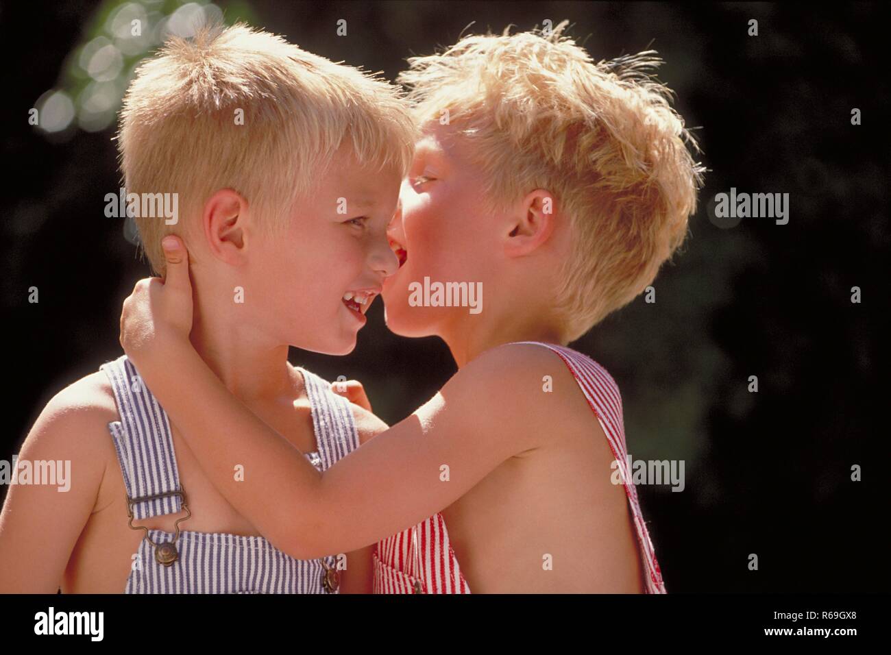 Portrait, Nahaufnahme, zwei 4 Jahre alte Zwillings-Jungen mit kurzen blonden Haaren bekleidet mit blau und rot-weiss gestreiften Latzhosen unterhalten sich in inniger Umarmung Stock Photo