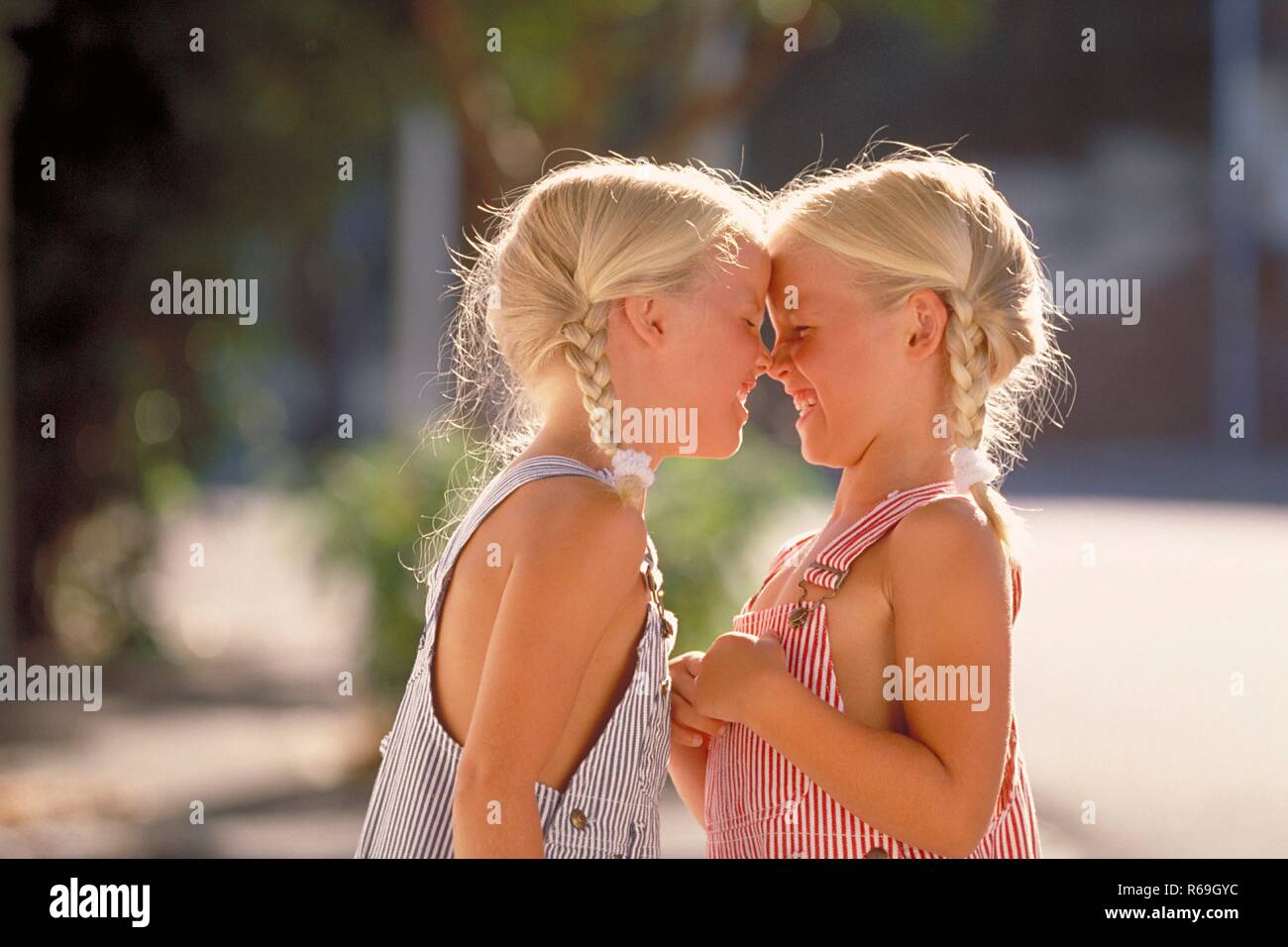 Portrait, Halbfigur, Profil von zwei lachenden 5 Jahre alten  Zwillings-Maedchen mit blonden geflochtenen Zoepfen bekleidet mit blau und rot-weiss gestreiften Latzhosen, die die Nasen aneinander reiben Stock Photo