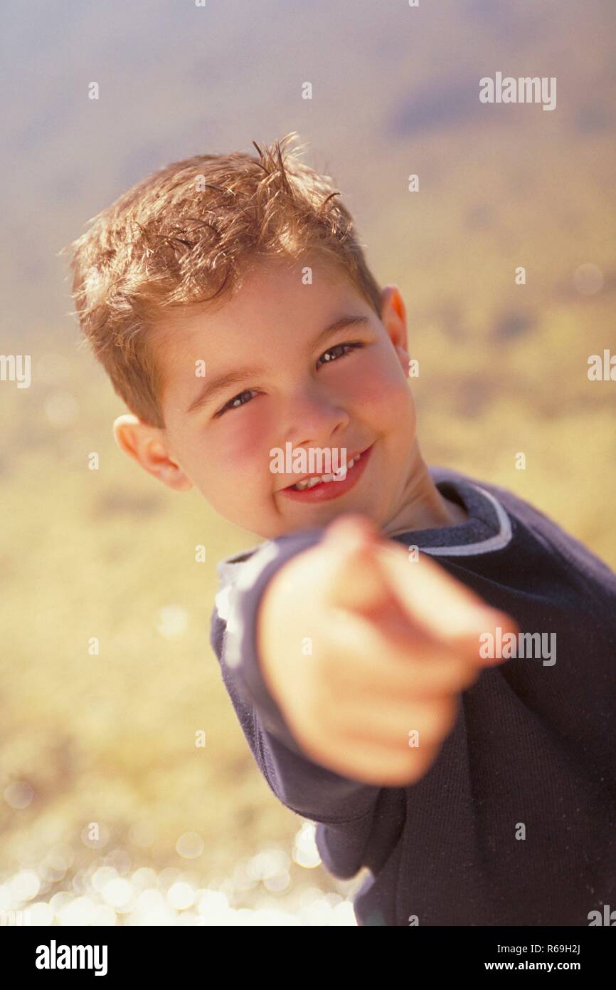 Strandszene, Portrait, 10-jaehriger Junge, bekleidet mit dunklem Hemd, steht vor einer glitzernden Wasserflaeche und zeigt mit dem Finger auf den Betrachter Stock Photo