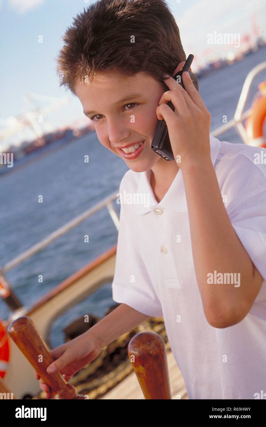 Hafen, Portrait, 14-jaehriger Junge bekleidet mit weissem Hemd steht auf See am Steuerrad eines Boots und telefoniert mit seinem Handy Stock Photo