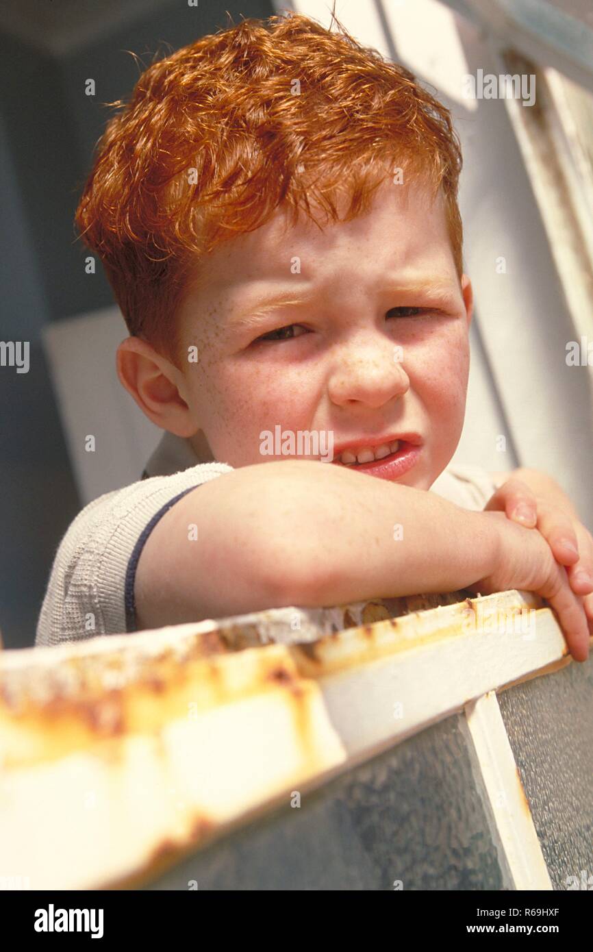 Portrait, Nahaufnahme, rothaariger Junge mit Sommersprossen, 6 Jahre, bekleidet mit weissem Hemd und blauer Latzhose schaut traurig aus einem Fenster mit rostigen Rahmen heraus Stock Photo