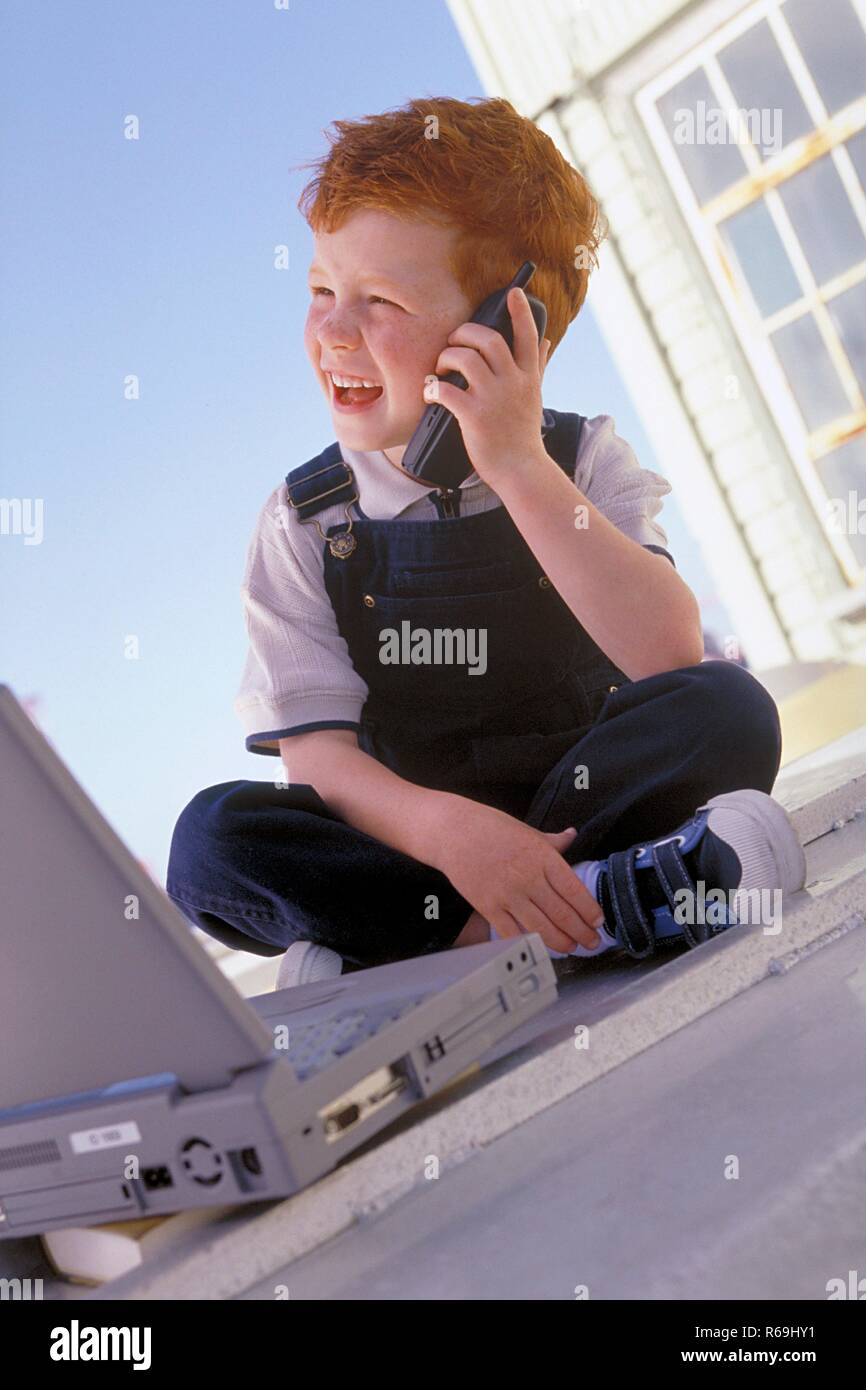 Portrait, 5-jaehriger rothaariger Junge mit Sommersprossen bekleidet mit blauer Latzhose sitzt mit Handy und Laptop am Fenster mit Sicht auf blauen Himmel Stock Photo