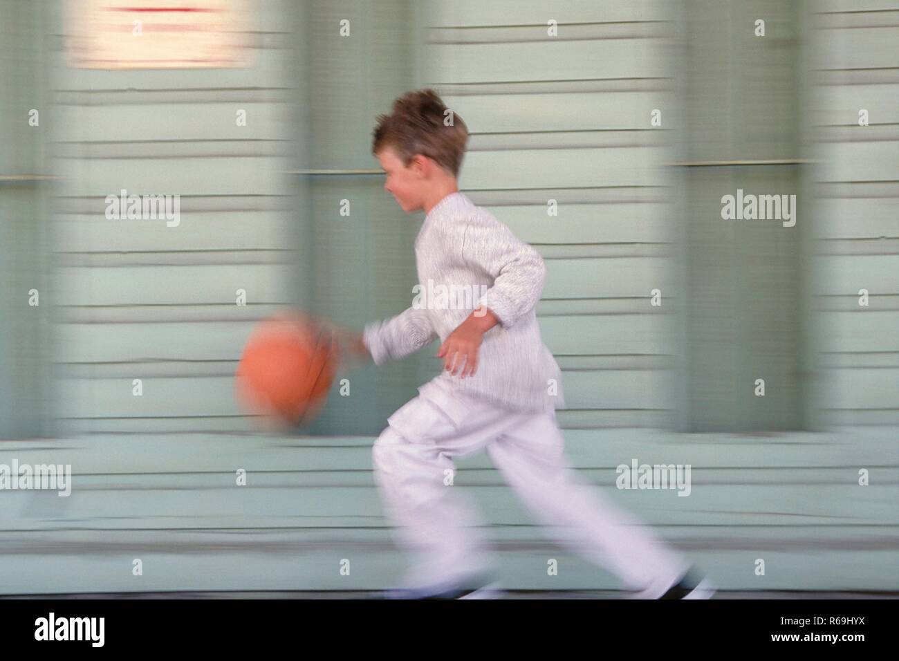 Portrait, Ganzfigur, 8- jaehriger Junge bekleidet mit grauer Hose, Pullover und Turnschuhen laeuft mit seinem orangem Basketball an einer graublau gestrichenen Fassade vorbei Stock Photo