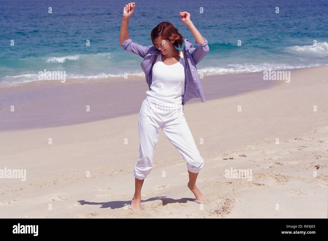 Strandszene, Portrait, Ganzfigur, junge Frau mit glatten braunen Haaren, bekleidet mit weisser Hose, T-Shirt und blauem Hemd tanzt barfuss am Strand Stock Photo