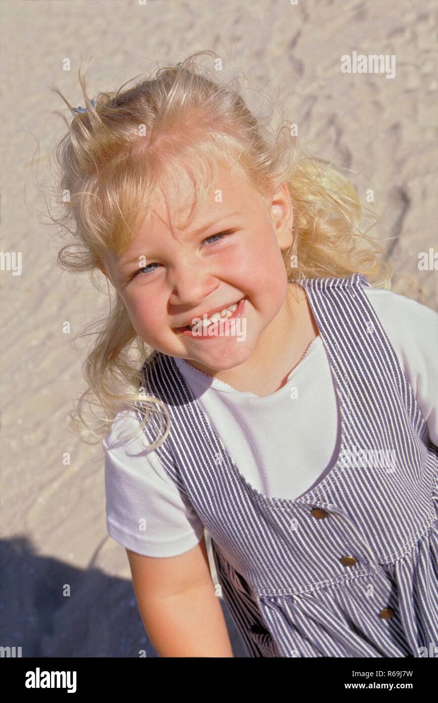 Strandszene, Portrait, Halbfigur, blond gelocktes Maedchen mit blauen Augen, 4 Jahre, bekleidet mit weissem T-Shirt und blau-weiss gestreiftem Kleid, zeigt lachend ihre Milchzaehne Stock Photo