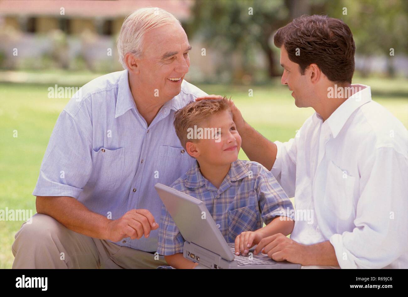 Parkszene, Portrait, 3 Generationen, weisshaariger Senior, dunkelhaariger Mann, Mitte 30, bekleidet mit hellen Hemden und blonder Junge mit kariertem Hemd, 6 Jahre, sitzen mit einem Laptop auf einer Wiese Stock Photo