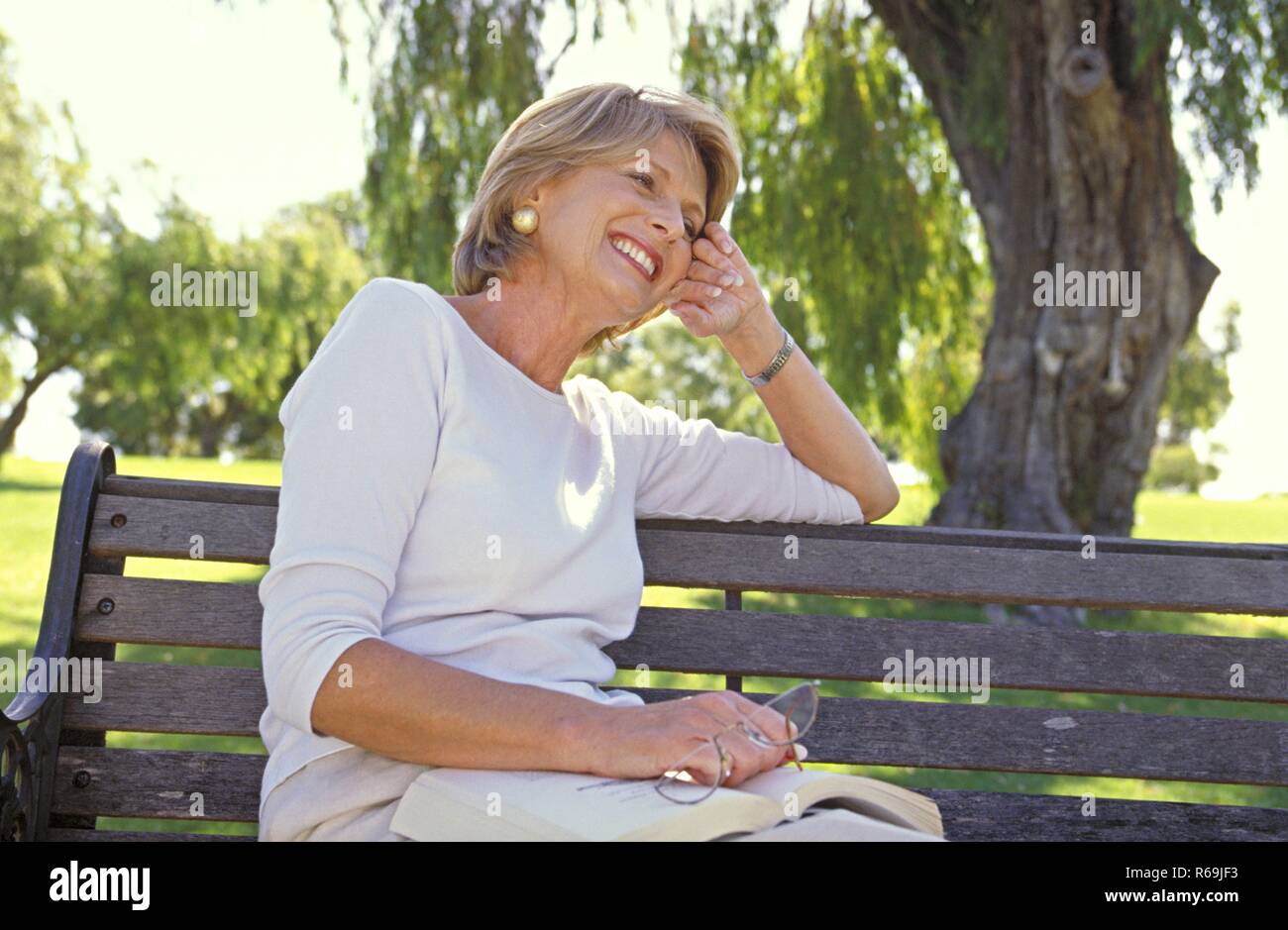 Portrait, Parkszene, blonde Frau, Mitte 50, bekleidet mit heller Hose und Top, sitzt entspannt mit einem Buch und ihrer Lesebrille in der Hand auf einer Parkbank unter einem Baum Stock Photo