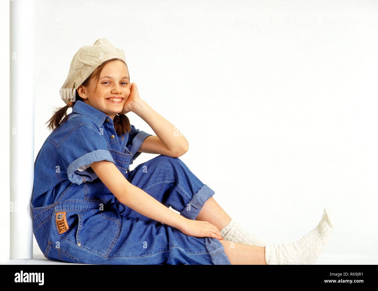 Ganzfigur, Maedchen, 11 Jahre, braune Zoepfe, bekleidet mit Kappe, Jeans  Hemd und Latzhose und weissen Socken, sitzt grinsend an eine weisse Saeule  gelehnt Stock Photo - Alamy