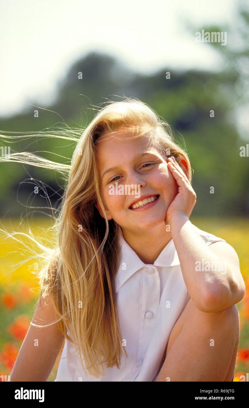 Portrait, laechelndes Maedchen mit langen braunen Haaren, 13 Jahre,  bekleidet mit weisser aermellosen Bluse und Shorts sitzt auf einer mit  gelben Blueten uebersaeten Wiese Stock Photo - Alamy