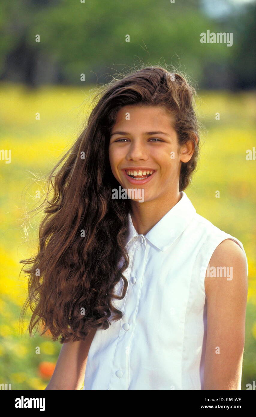 Portrait, laechelndes Maedchen mit langen braunen Haaren, 13 Jahre, bekleidet mit weisser aermellosen Bluse und Shorts sitzt auf einer mit gelben Blueten uebersaeten Wiese Stock Photo