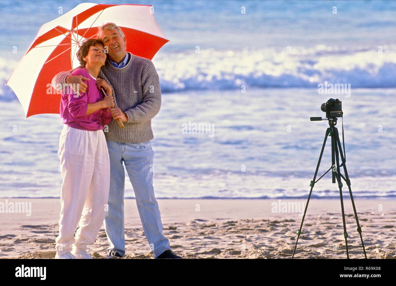 Seniorenpaar, Mann mit Jeans und Pullover und Frau mit weisser Hose und himbeerfarbener Strickjacke und rot-weissem Sonnenschirm machen am Strand ein Foto per Selbstausloeser Stock Photo