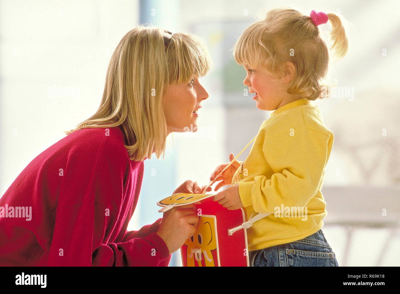 Mutter mit blonden Haar kauert vor ihrer blond bezopften Tochter, 4 Jahre, die eine grosse rot-gelbe Kindergartentasche traegt und spricht beruhigend auf sie ein Stock Photo