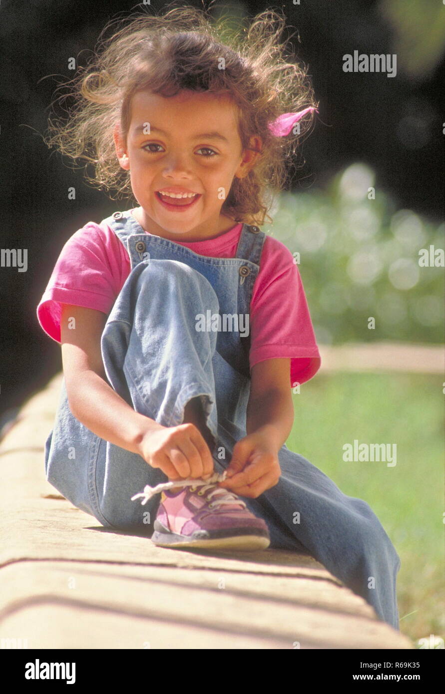 Parkszene, Ganzfigur, Maedchen mit langen braunen Haaren, 6 Jahre, bekleidet mit Jeans und pink farbenem T-Shirt, sitzt auf einer Steinmauer und bindet sich ihren Schuh zu Stock Photo