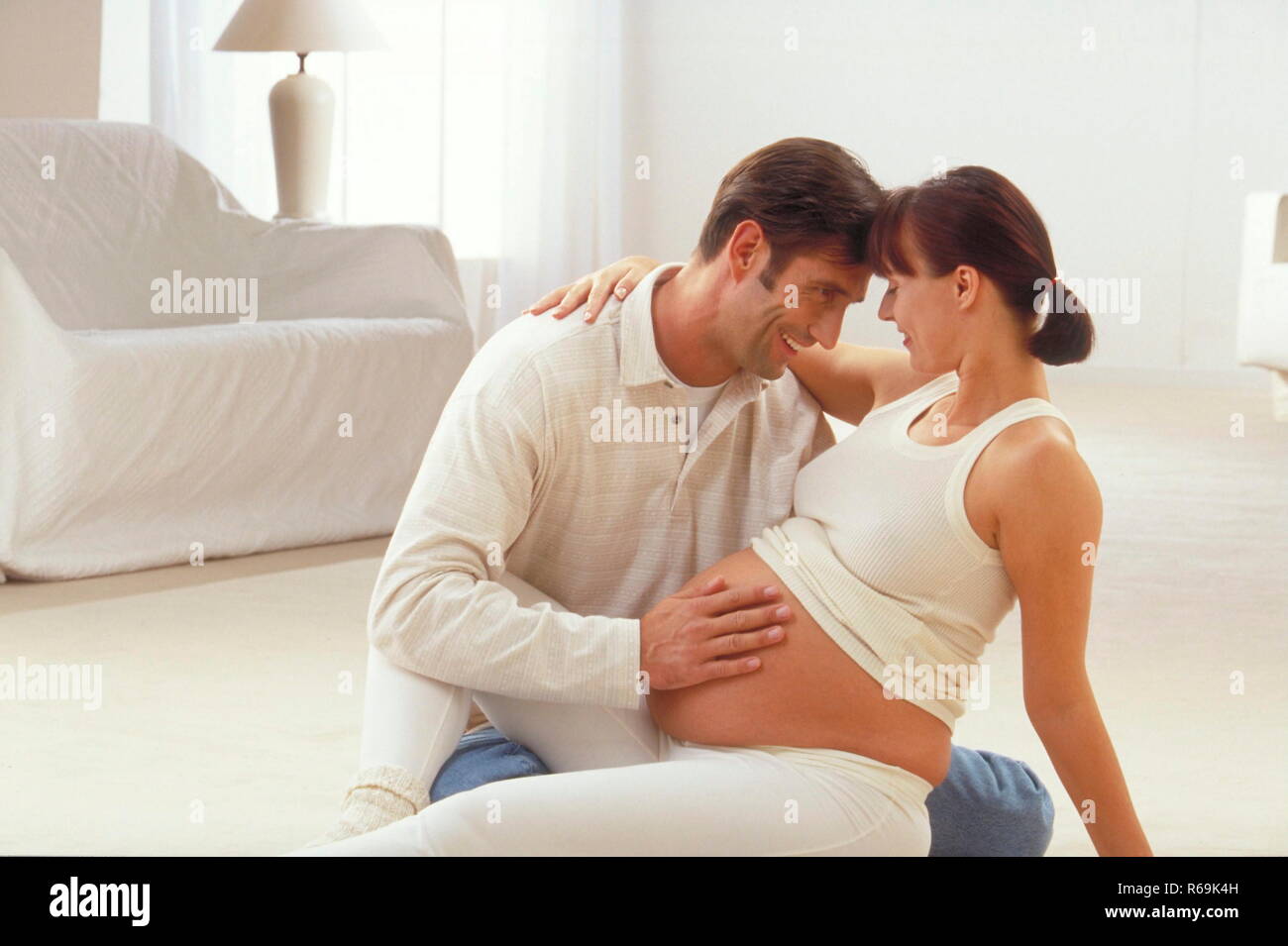 Portrait, Ehepaar sitzt im Wohnzimmer auf dem Teppichboden, der Mann streichelt den nackten Bauch seiner hochschwangeren Frau und fuehlt die Bewegungen seines Kindes Stock Photo