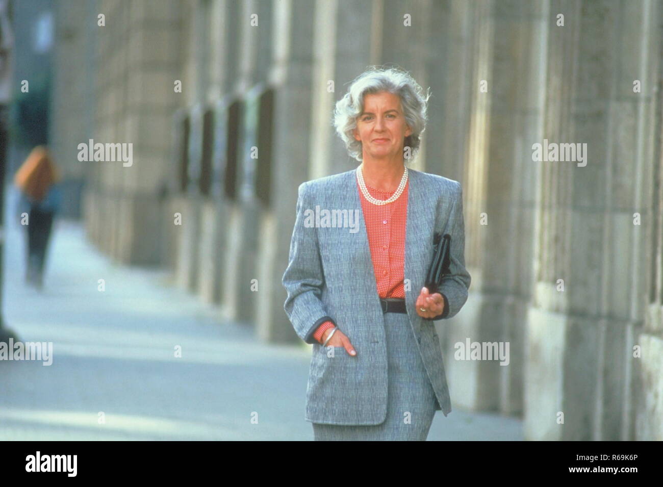 Portrait, Stadtszene, Frau, ca. 60 Jahre, bekleidet mit grauem Kostuem und Unterarmtasche geht auf dem Buergersteig an alten Haeuserfassaden vorbei Stock Photo