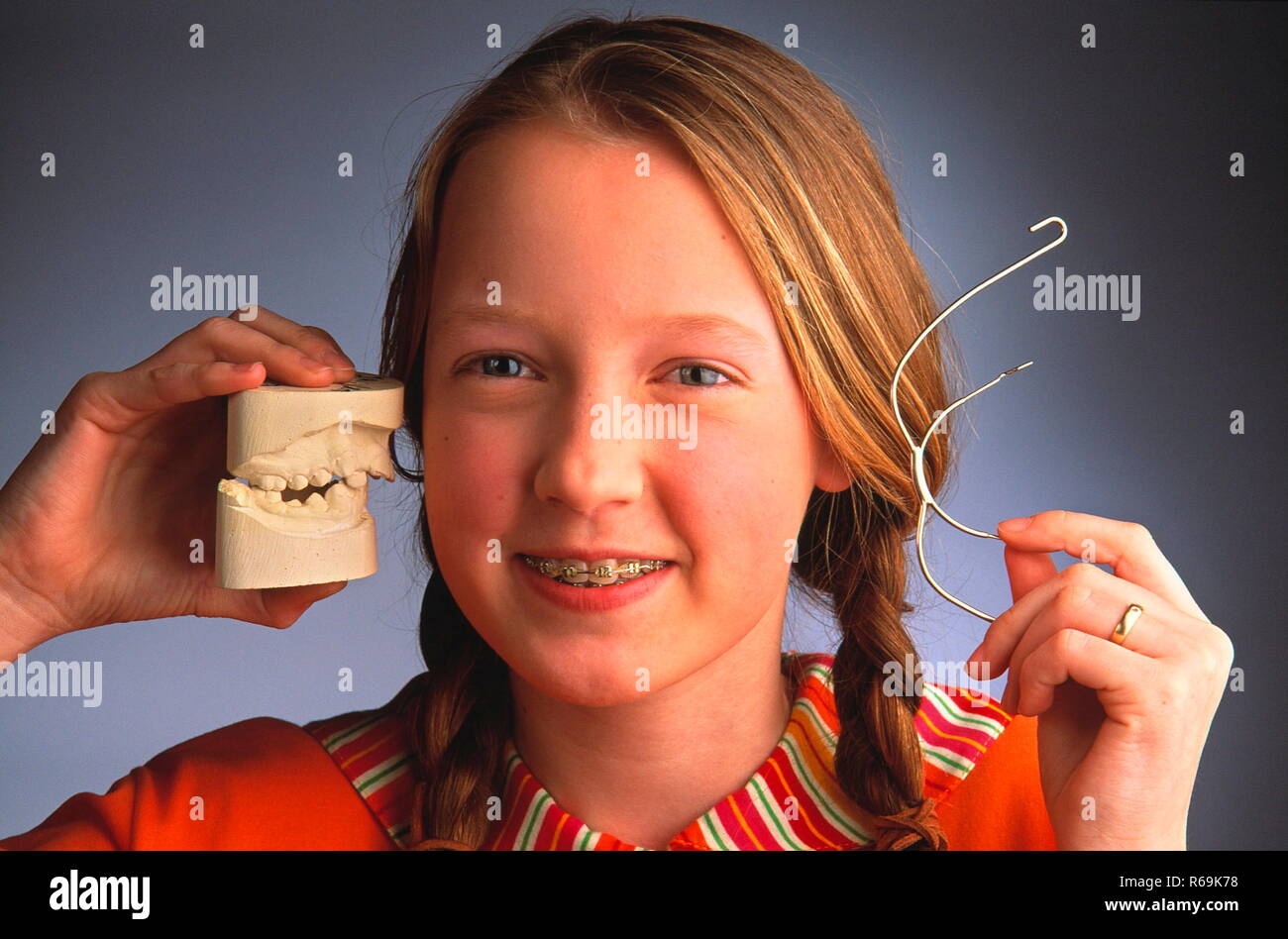 Portrait, Nahaufnahme, Maedchen mit Zoepfen, 13 Jahre, zeigt laechelnd ihre Zahnklammer und haelt den Abdruck ihres Gebisses und den Gesichtsbogen in den Haenden Stock Photo