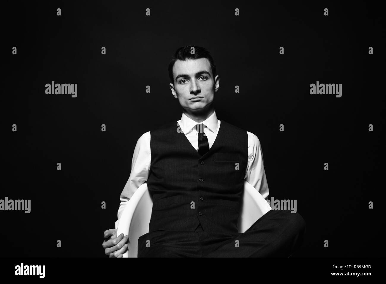Close up black and white portrait of a young man in a white shirt, black tie and vest, sitting and seriously looking at the camera, against plain stud Stock Photo