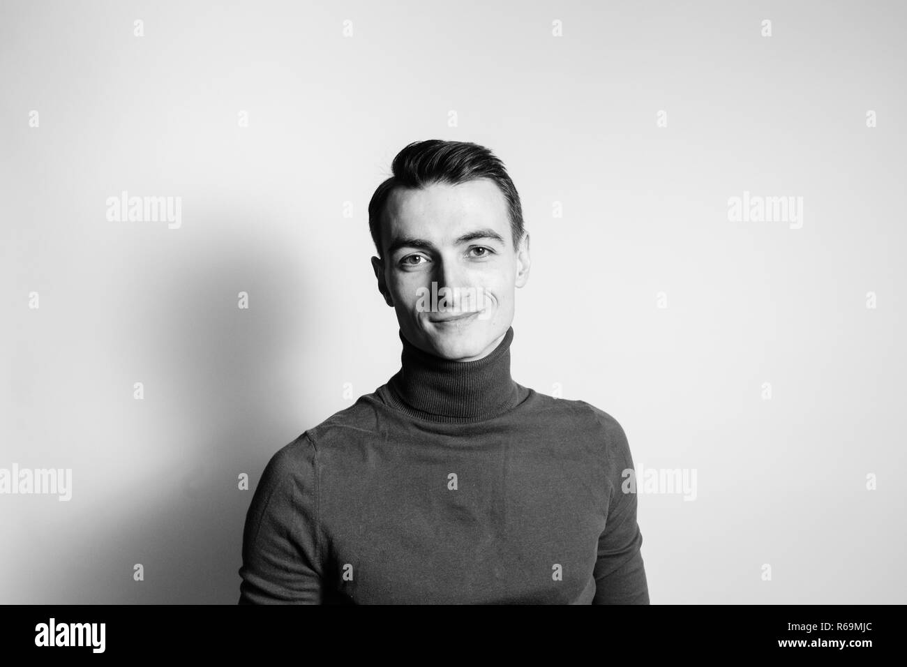 Close up black and white portrait of a young man wearing turtleneck jumper, smiling at the camera, against plain studio background Stock Photo