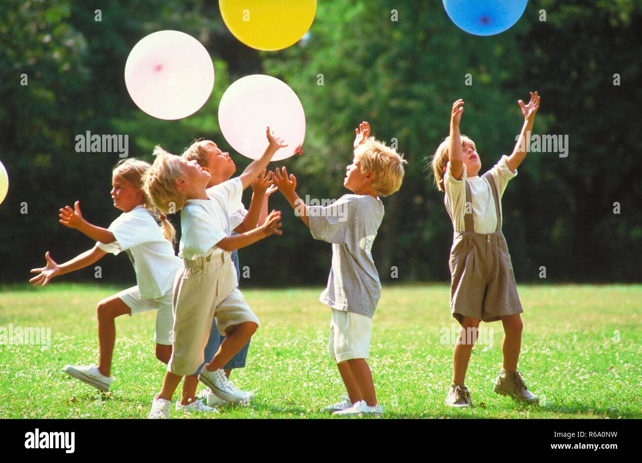 5 Kinder spielen auf einer mit Gaensebluemchen uebersaeaten Wiese im Park mit bunten Luftballons Stock Photo