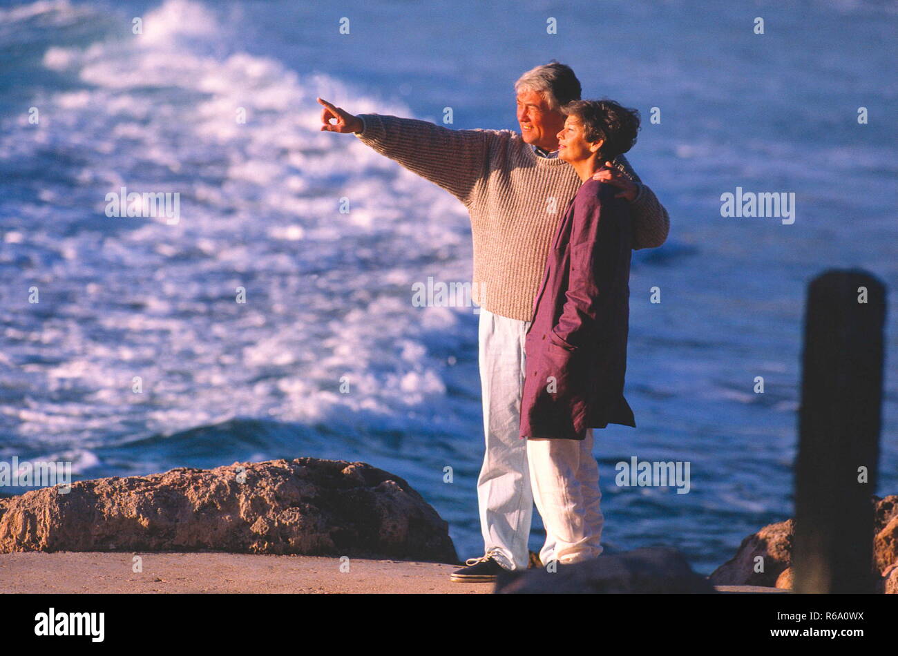 Portrait, Paar, ca. 50 Jahre, steht an Felskueste, in den Wellen spiegelt sich die Sonne Stock Photo