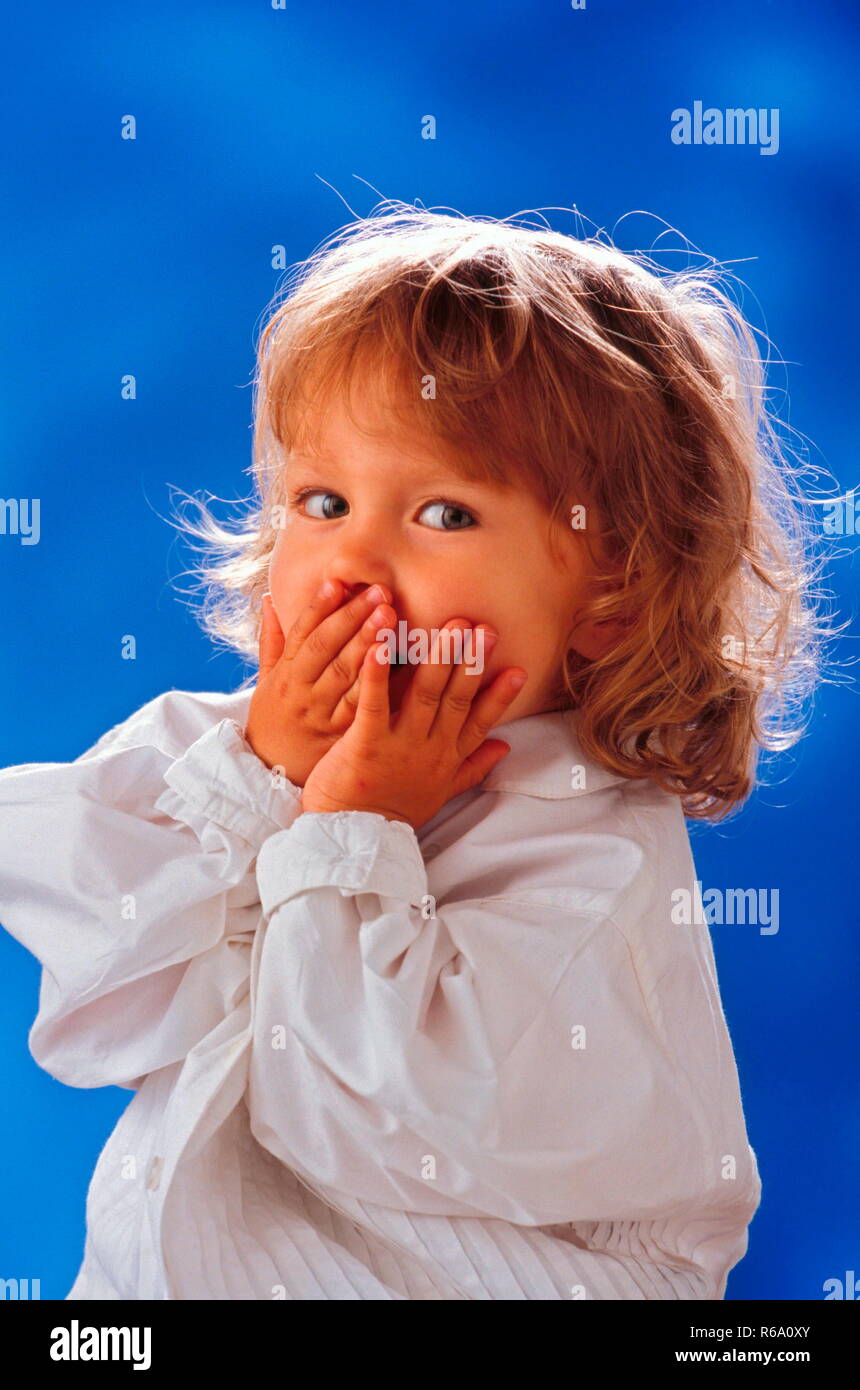 Portrait, Maedchen, 3 Jahre, in weisser Spitzenbluse, haelt sich die Haende vor den Mund, baluer Hintergrund Stock Photo