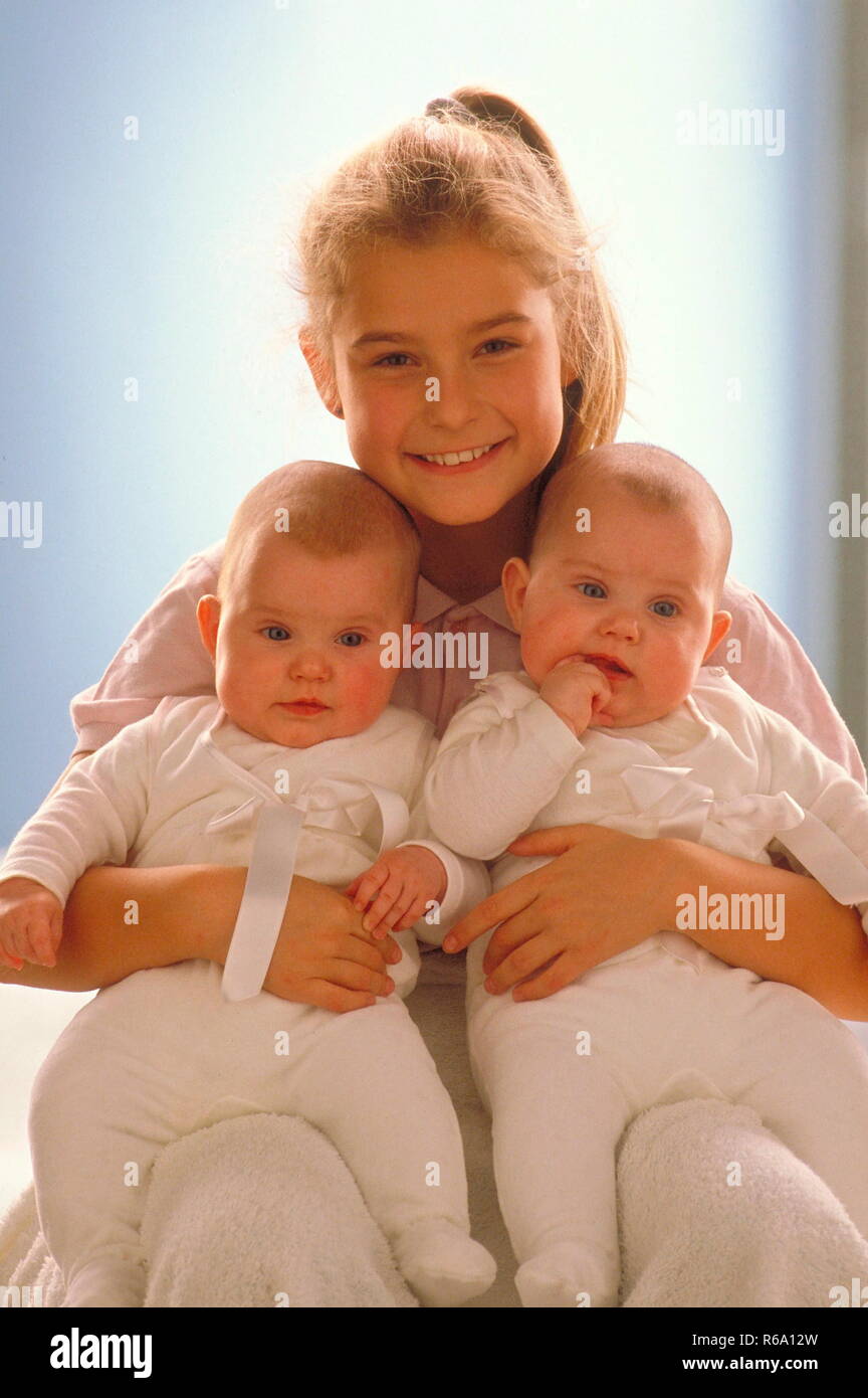 Portrait, sitzendes Maedchen mit Pferdeschwanz, 8 Jahre, die ihre beiden Zwillingsschwestern, ca. 6 Monate, in den Armen haelt Stock Photo