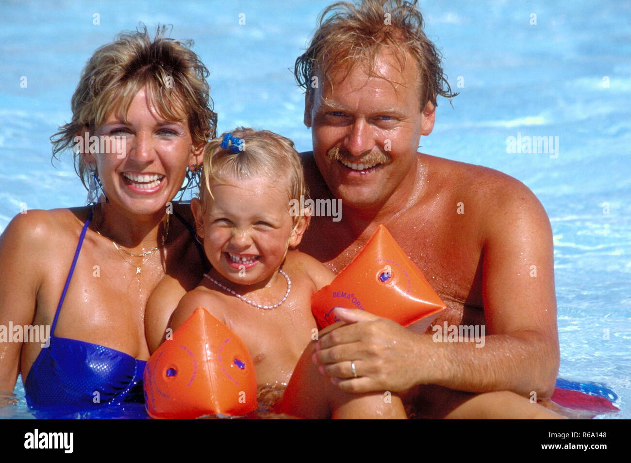 Portrait, blondes Paar sitzt mit Kind, 2 Jahre, mit orangen Schwimmfluegeln im Schwimmbecken Stock Photo
