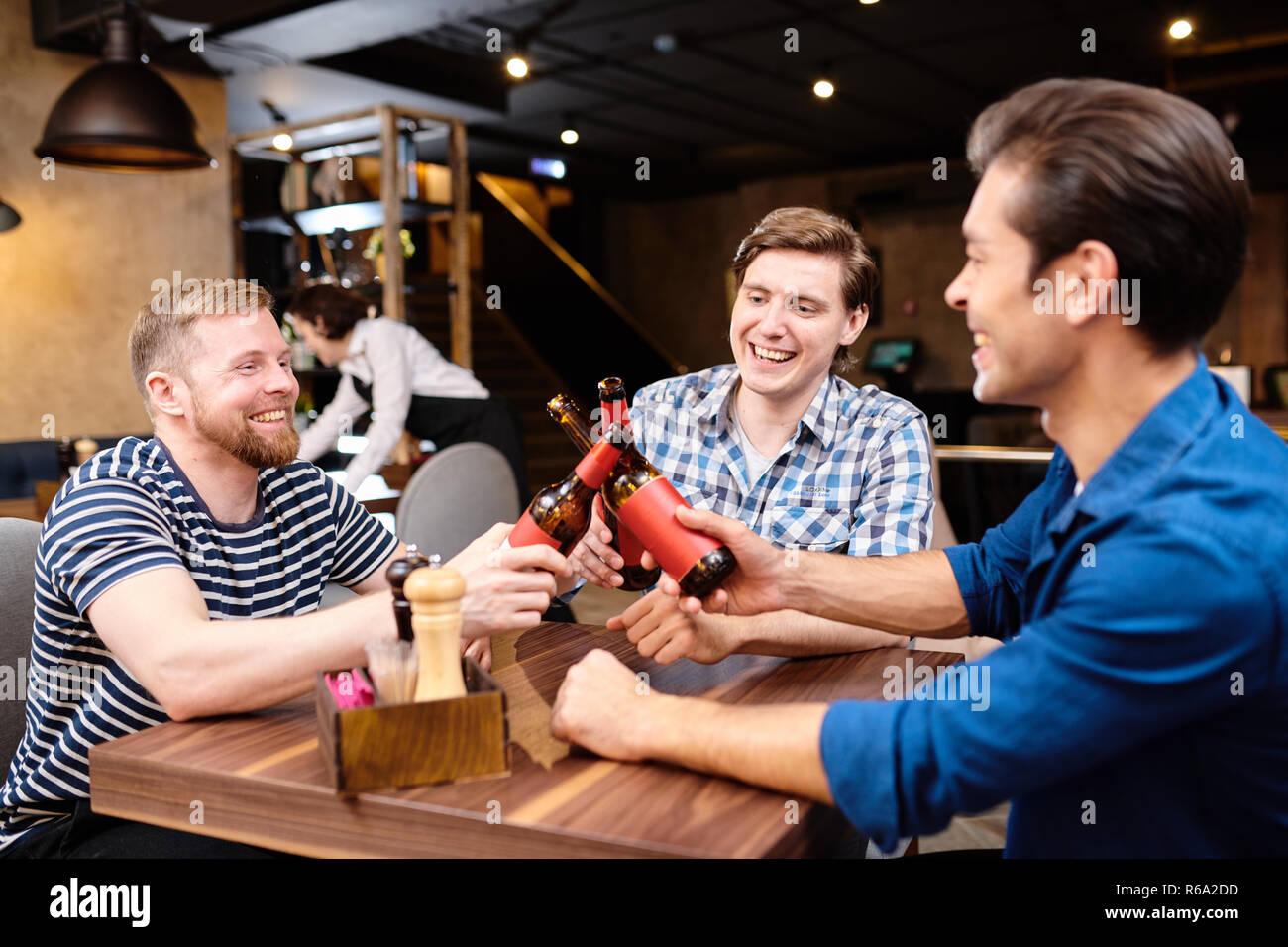 Group of carefree handsome young friends in casual outfit sitting at table  in pub and clinking beer bottles while drinking or gathering Stock Photo -  Alamy