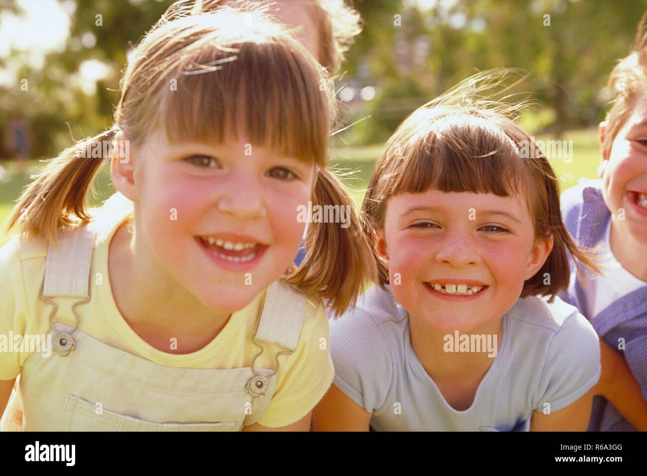 Parkszene, Portrait, Nahaufnahme, 2 dunkelblonde Maedchen, 6 Jahre alt, bekleidet mit hellgelben und hellblauen T-Shirtszeigen ihre Zaehne Stock Photo