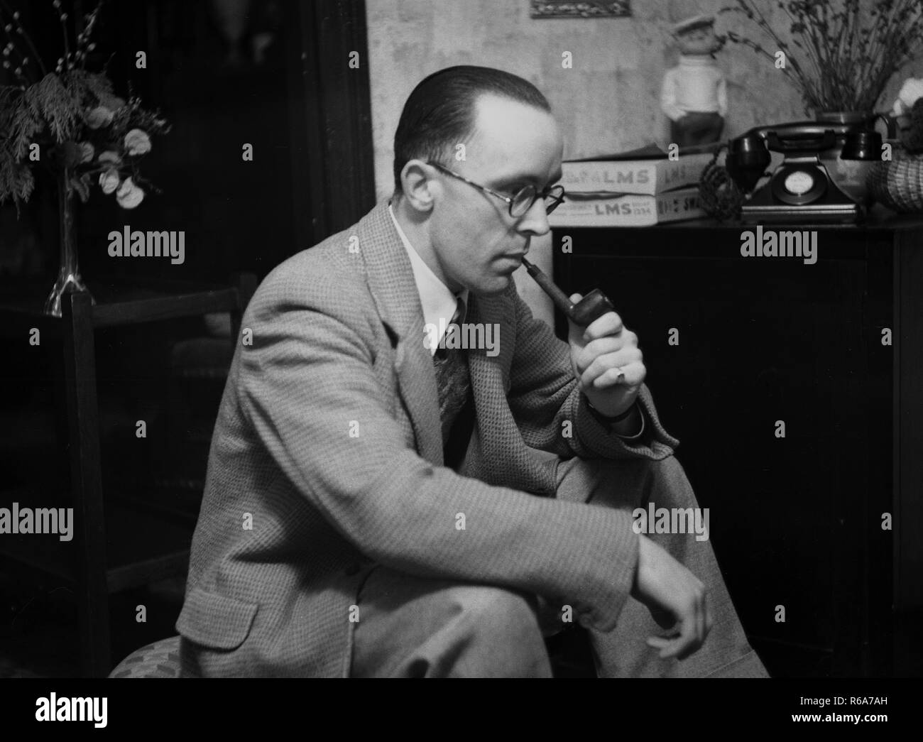 1950s, historical, a man in a suit sitting smoking a pipe indoors at home, England. Pipe smoking was popular at this time. Also seen in the picture a bakelite GPO telephone of the era. Stock Photo