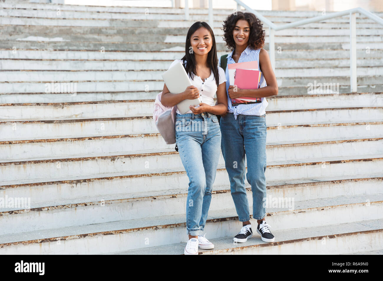 Full length of two happy young students girls standing outdoors ...