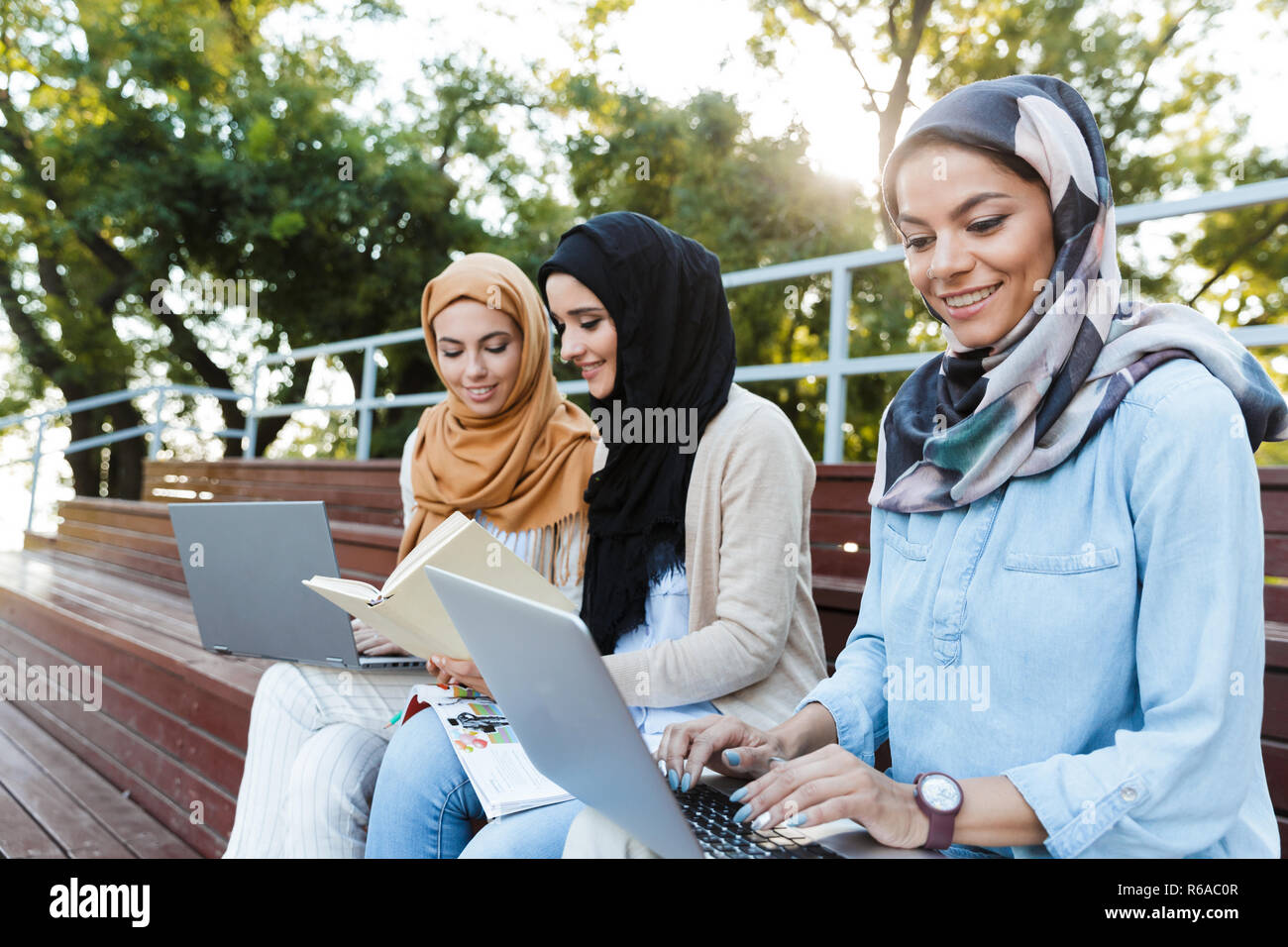 Photo of cheerful islamic women wearing headscarfs resting in green park and studying on laptop Stock Photo