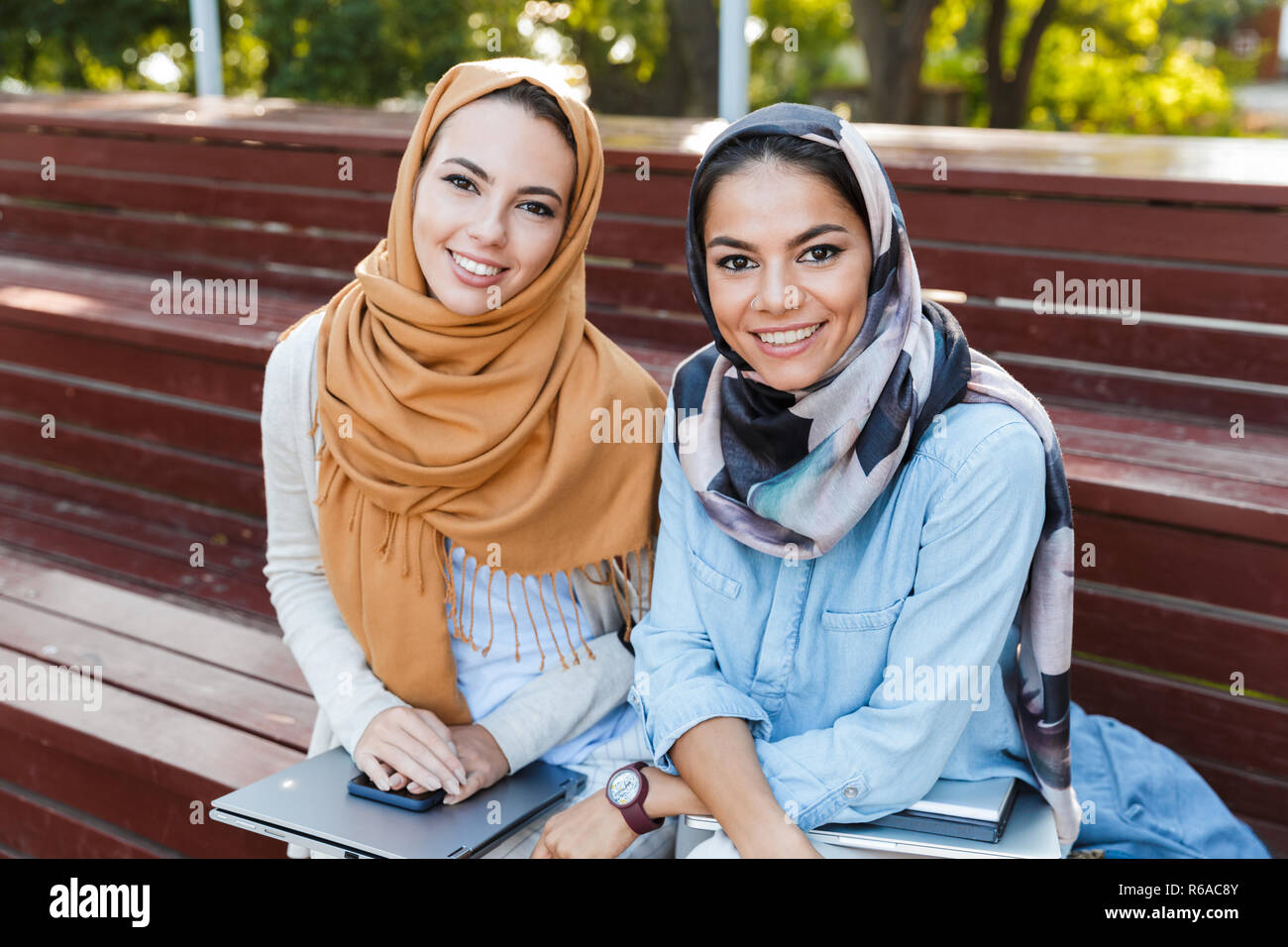 Photo of two muslim women wearing headscarfs smiling at camera while sitting on bench in park Stock Photo