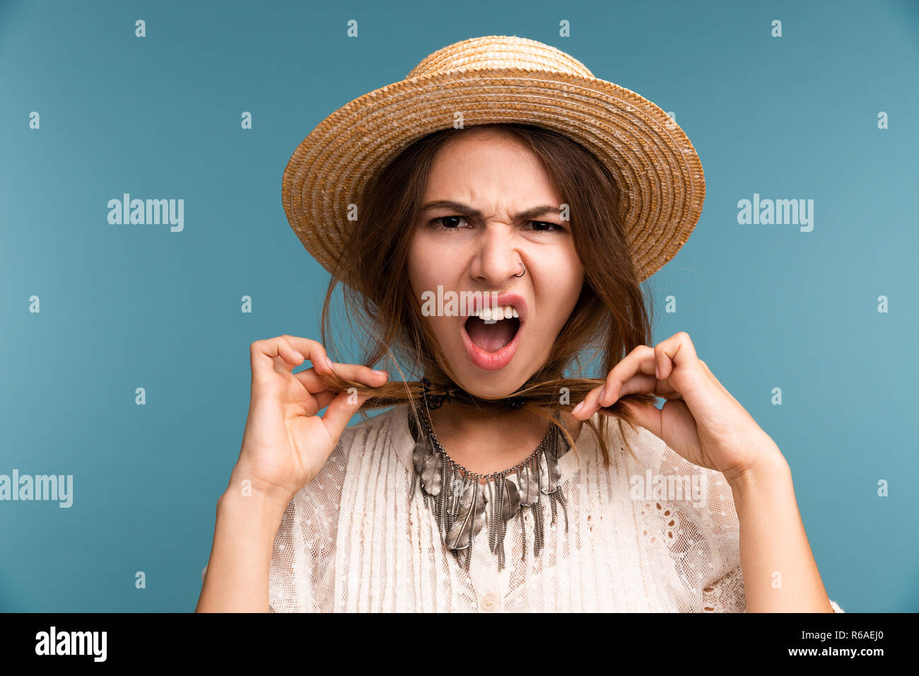 Portrait of a young angry girl in summer hat isolated over blue background, strangling herself with her hair Stock Photo