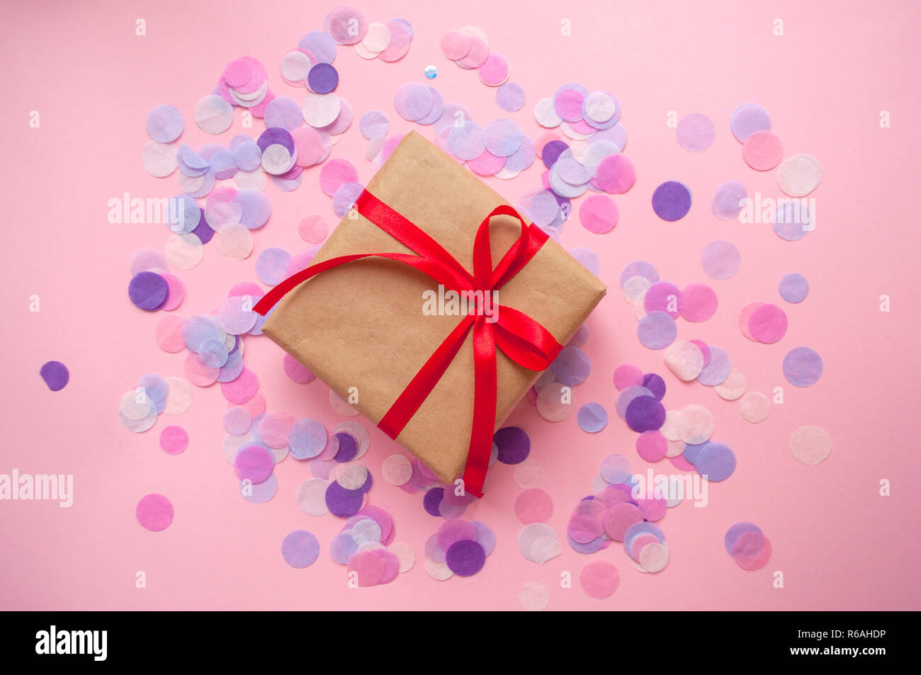 Gift box wrapped in brown colored craft paper and tied with red bow on pink background with colofrul confetti. Stock Photo