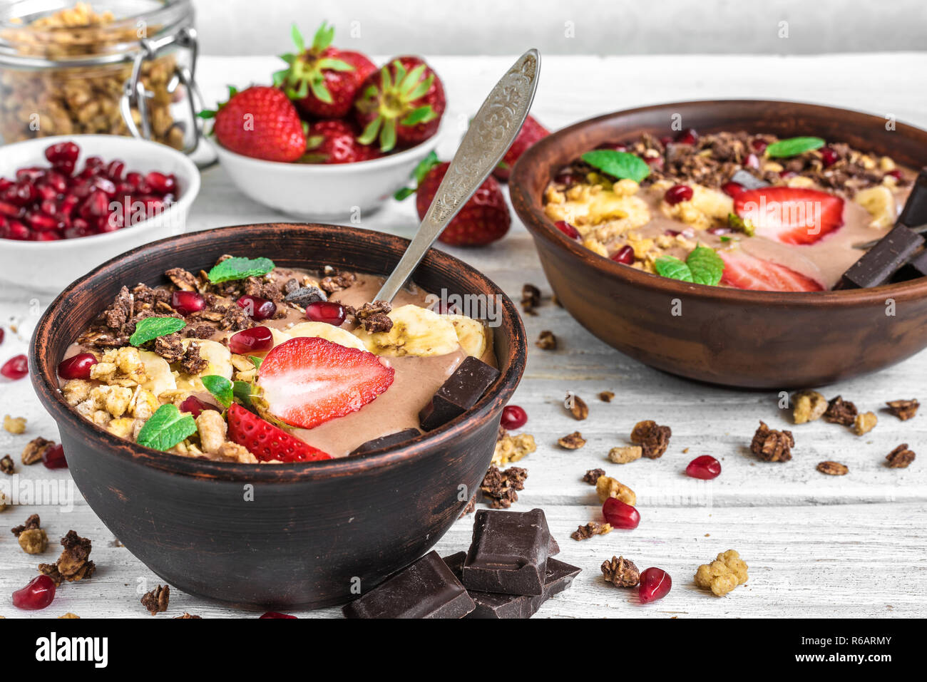 chocolate banana protein smoothie bowls with granola, strawberry, pomegranate and chocolate bars served for breakfast on white wooden table. close up. Stock Photo