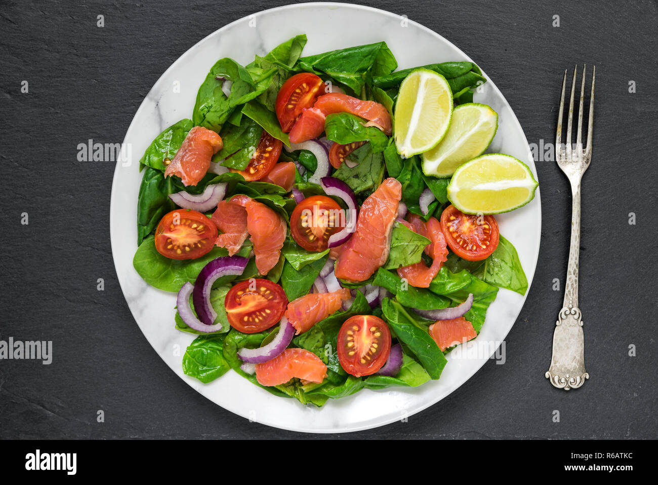 fresh salad with salmon, spinach, cherry tomatoes, red onion and basil in marble plate with fork over dark slate background. healthy food concept. top Stock Photo