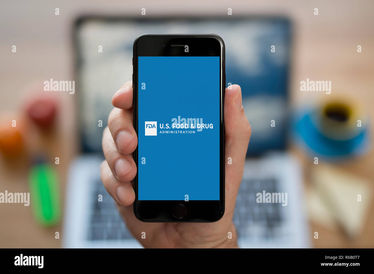 A man looks at his iPhone which displays the FDA (U S Food and Drug Administration) logo, while sat at his computer desk (Editorial use only). Stock Photo