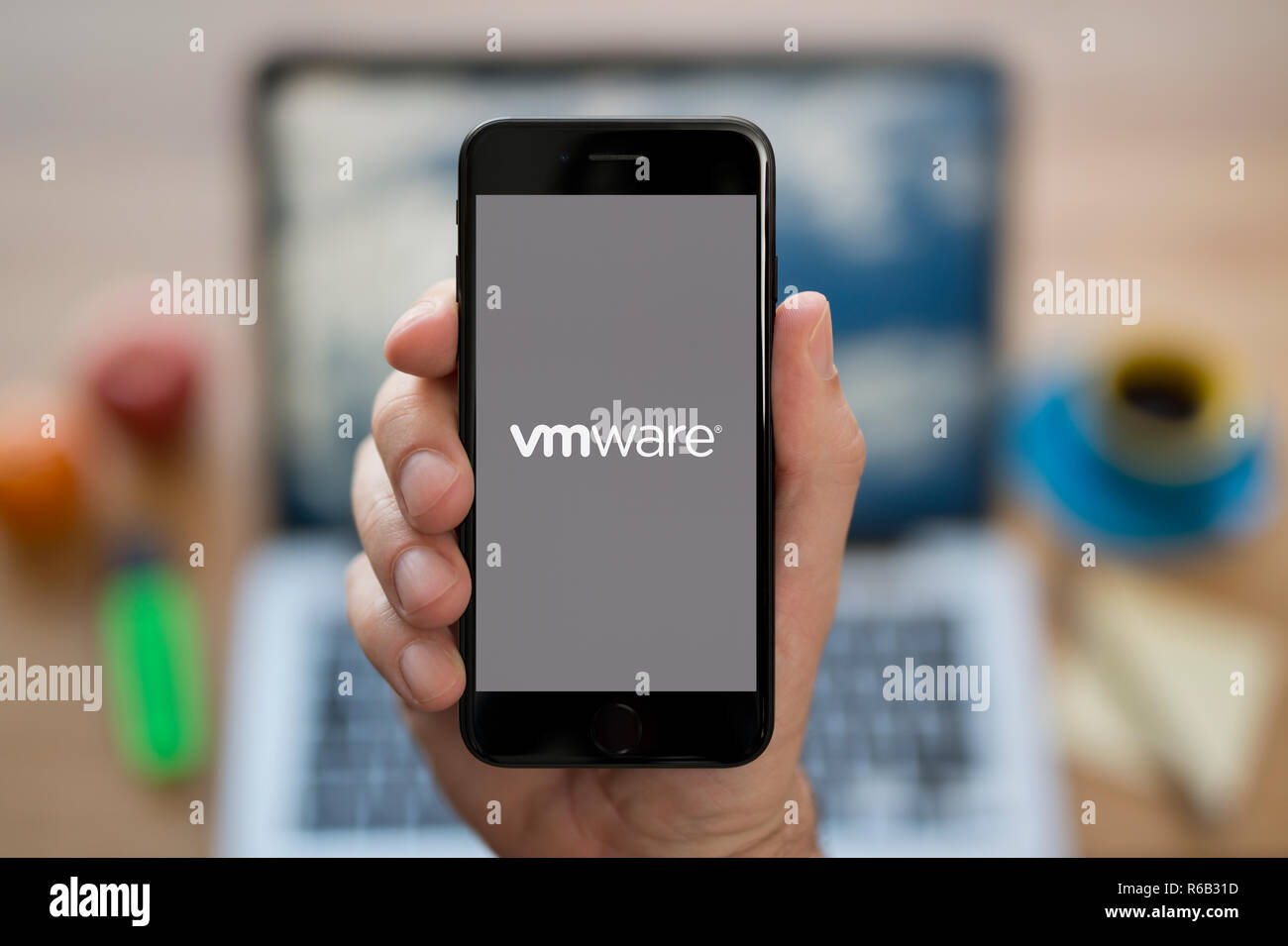 A man looks at his iPhone which displays the VMWare logo, while sat at his computer desk (Editorial use only). Stock Photo