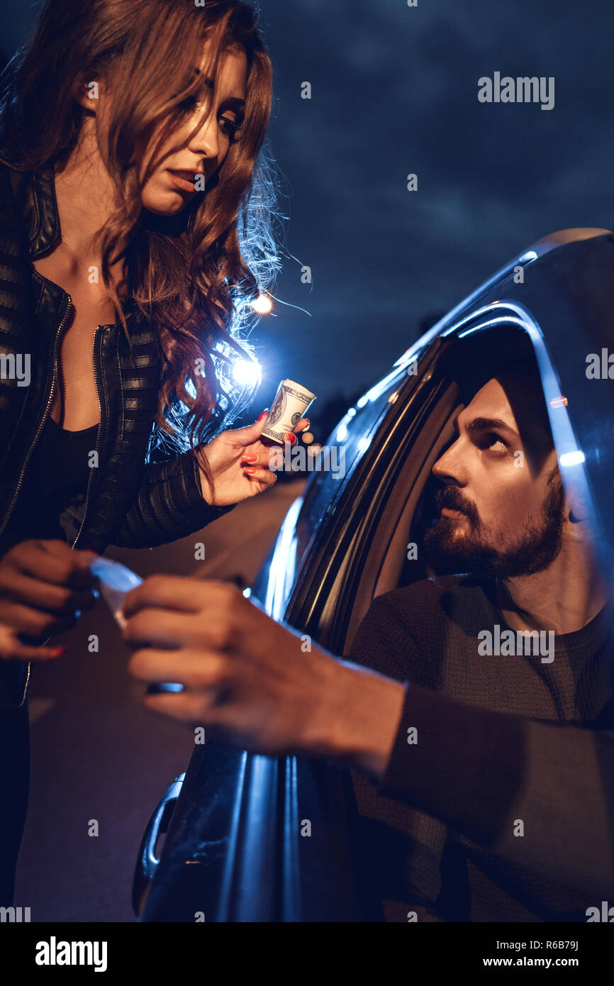 The dealer sells a little bag of heroin or cocaine to a young woman from a car, and she gives him money. Stock Photo