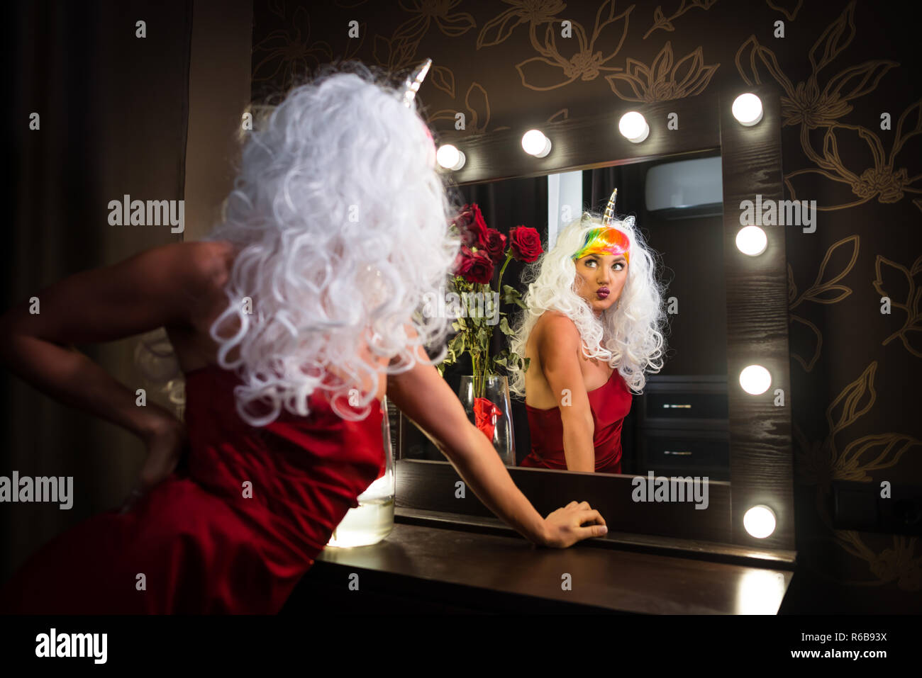 Funny young woman in unusual wig looking at himself in mirror in dressing room with flowers. Strange lady in red dress. Girl unicorn makes faces. Stock Photo