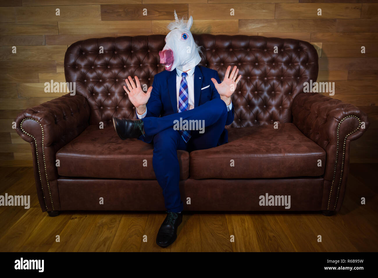 Funny unicorn in elegant suit siting on sofa like a boss and showing do not bother me gesture. Portrait of unusual man at home. Freaky guy in mask. Stock Photo