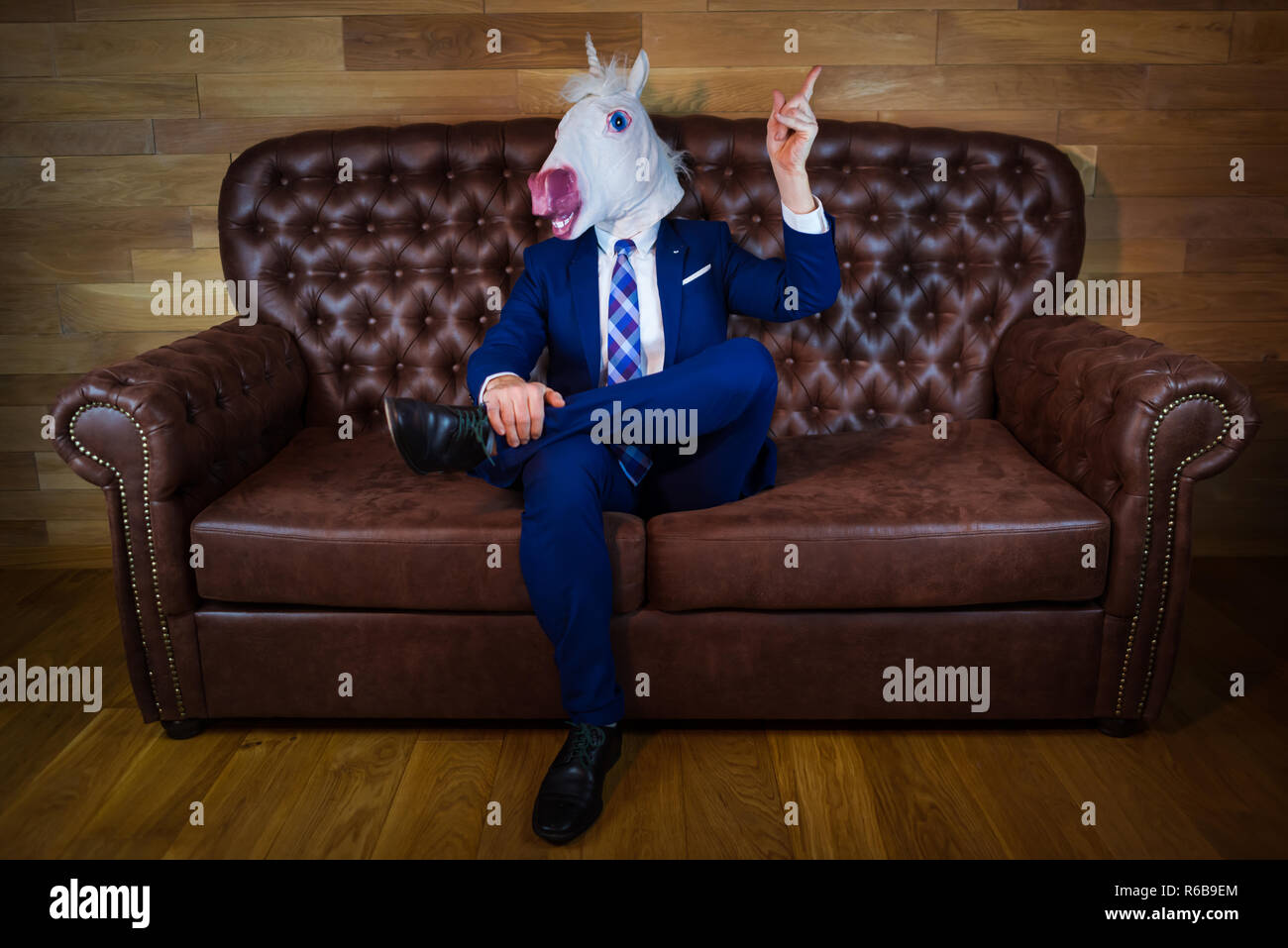 Funny unicorn in elegant suit siting on sofa like a boss and showing hand gesture with raised index finger. Unusual man at home. Freaky guy in mask. Stock Photo