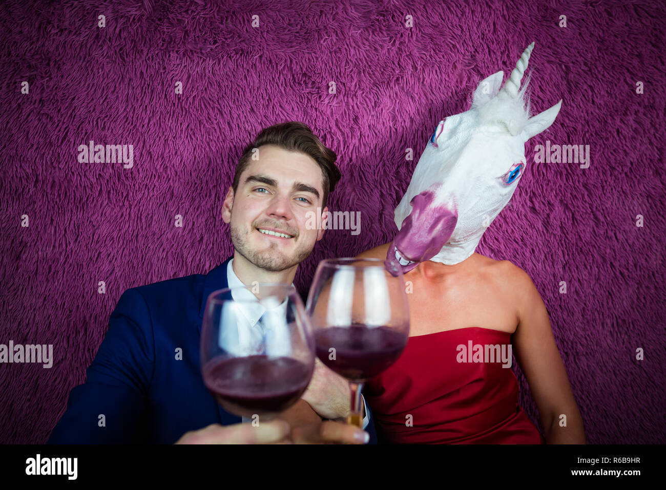 Portrait of strange couple on purple background. Freaky young woman in comical mask drinking wine with guy in suit. Young man with girl unicorn. Stock Photo