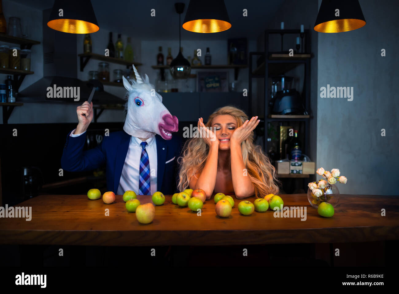 Funny couple playing at bar counter in kitchen with apples on table. Blonde girl have fun with boyfriend in mask. Unicorn in suit with young woman Stock Photo