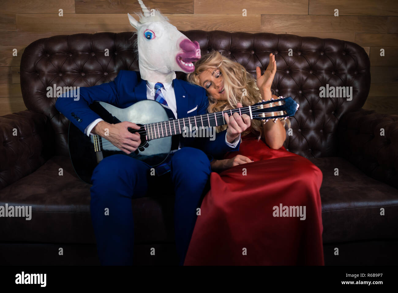 Man in suit and mask playing music on guitar for pretty girl in red dress. Unusual couple on leather sofa in stylish room. Unicorn with girlfriend Stock Photo