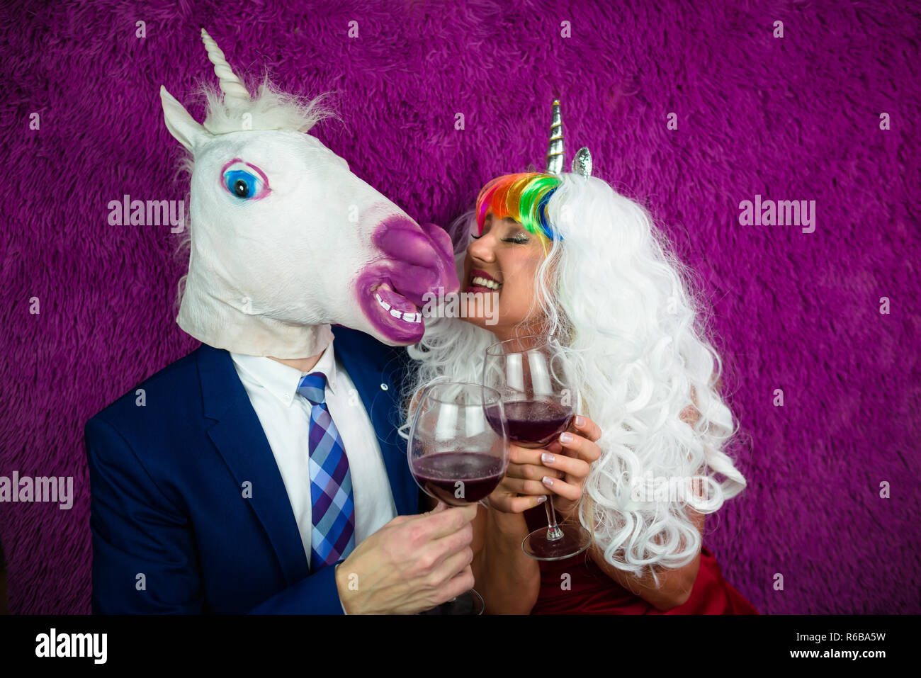 Portrait of strange couple on purple background. Freaky young woman in unusual wig drinking wine with man in mask and suit. Unicorn with girlfriend. Stock Photo