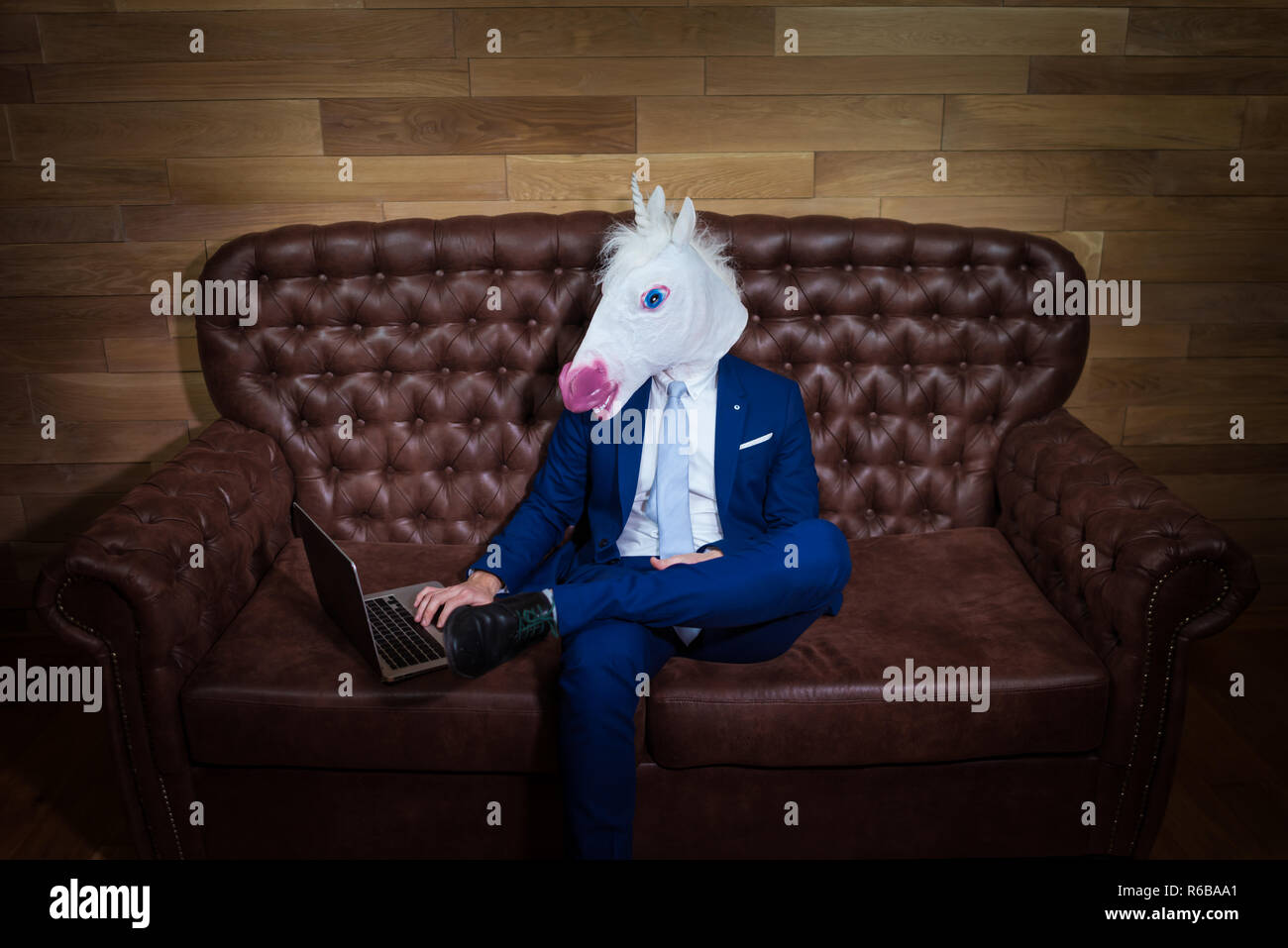 Strange guy in elegant suit working in home office. Unusual young man in mask on background of wooden wall. Funny unicorn sits on sofa like a boss. Stock Photo